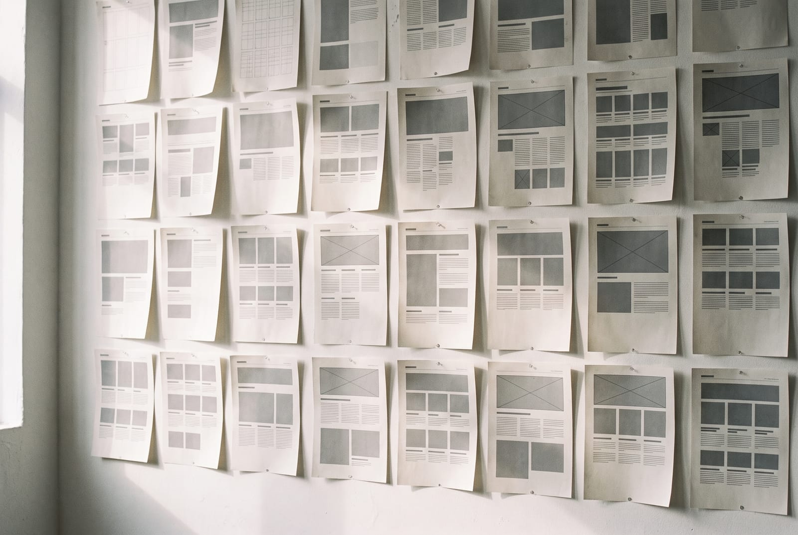 A studio wall of printed layout sheets and typographic grids pinned in a strict grid, lit from the side.